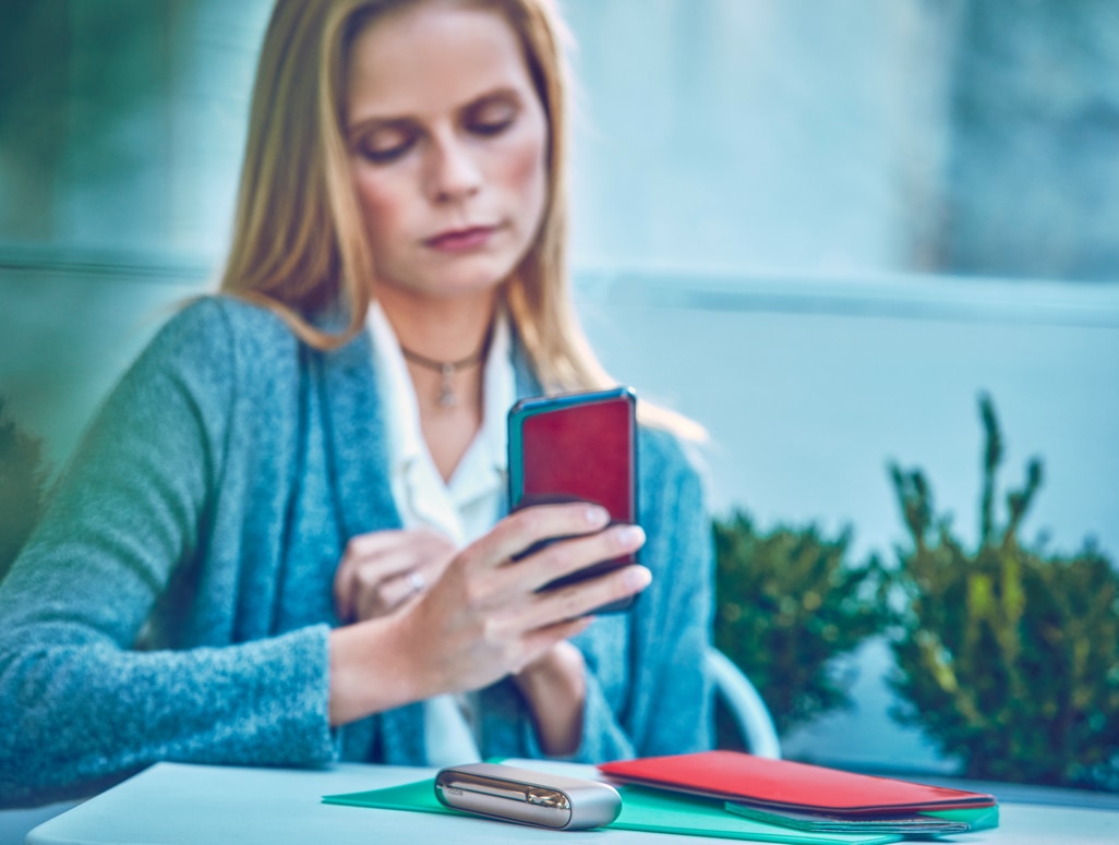 Mujer rubia mirando el celular con un dispositivo IQOS sobre la mesa