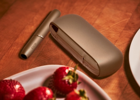 Soporte y cargador IQOS 3 DUO dorado brillante junto a un plato de fresas