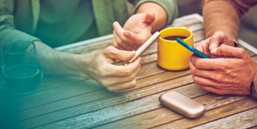 Two people talking at a table bar, one smokes IQOS and the other uses his phone