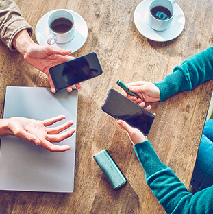 Dos personas con teléfonos móviles sentadas a la mesa; una de ellas también sostiene un dispositivo IQOS ILUMA PRIME color Jade Blue.