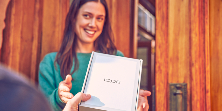 Persona entregándole la caja de un dispositivo IQOS a una mujer sonriente.